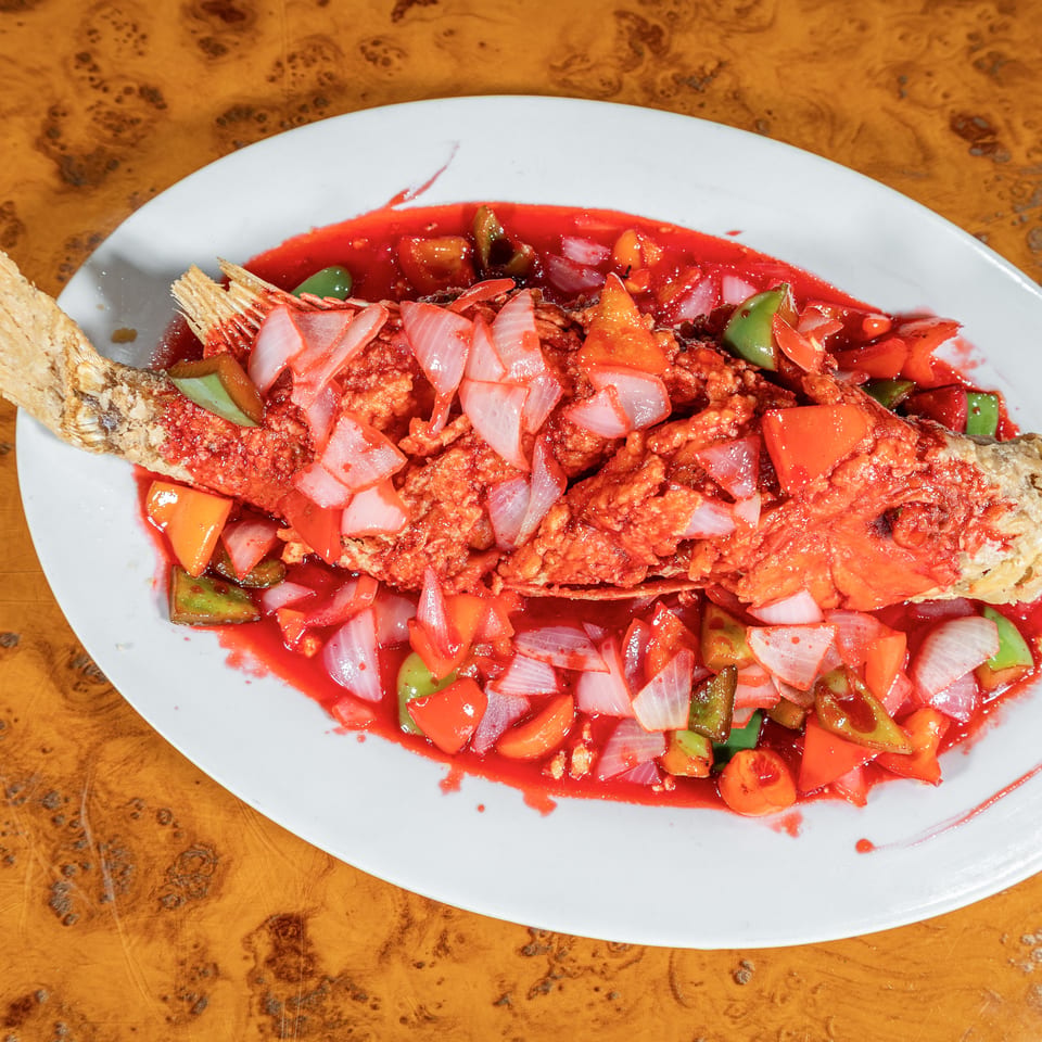 Whole fried fish topped with onions, tomatoes, and green peppers in bright red sauce on a white oval plate.