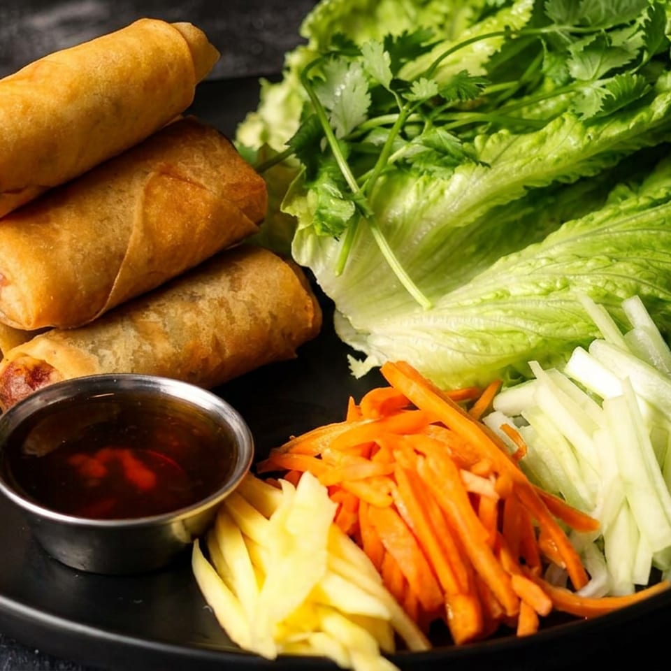Crispy spring rolls with dipping sauce, lettuce, shredded carrots, cucumber, and pineapple arranged on a black plate.
