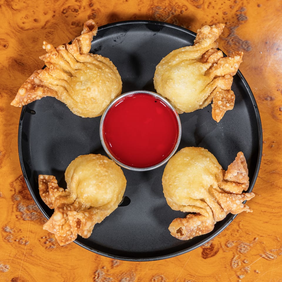 Four golden fried dumplings surround a bowl of bright red dipping sauce on a black plate atop a wooden table.
