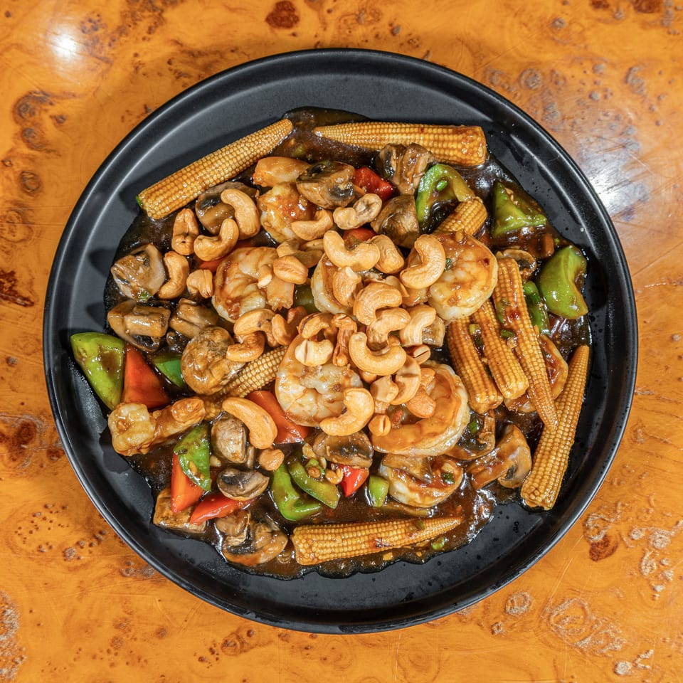 Black plate of stir-fried shrimp, cashews, mushrooms, baby corn, and colorful peppers in savory sauce.