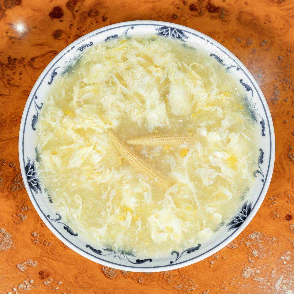 Bowl of Chinese egg drop soup with baby corn in a decorative porcelain bowl on a wooden table.