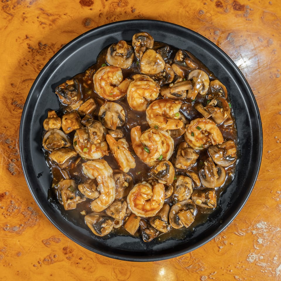 Black plate filled with shrimp and sliced mushrooms in a glossy brown sauce on a wood-grain table.
