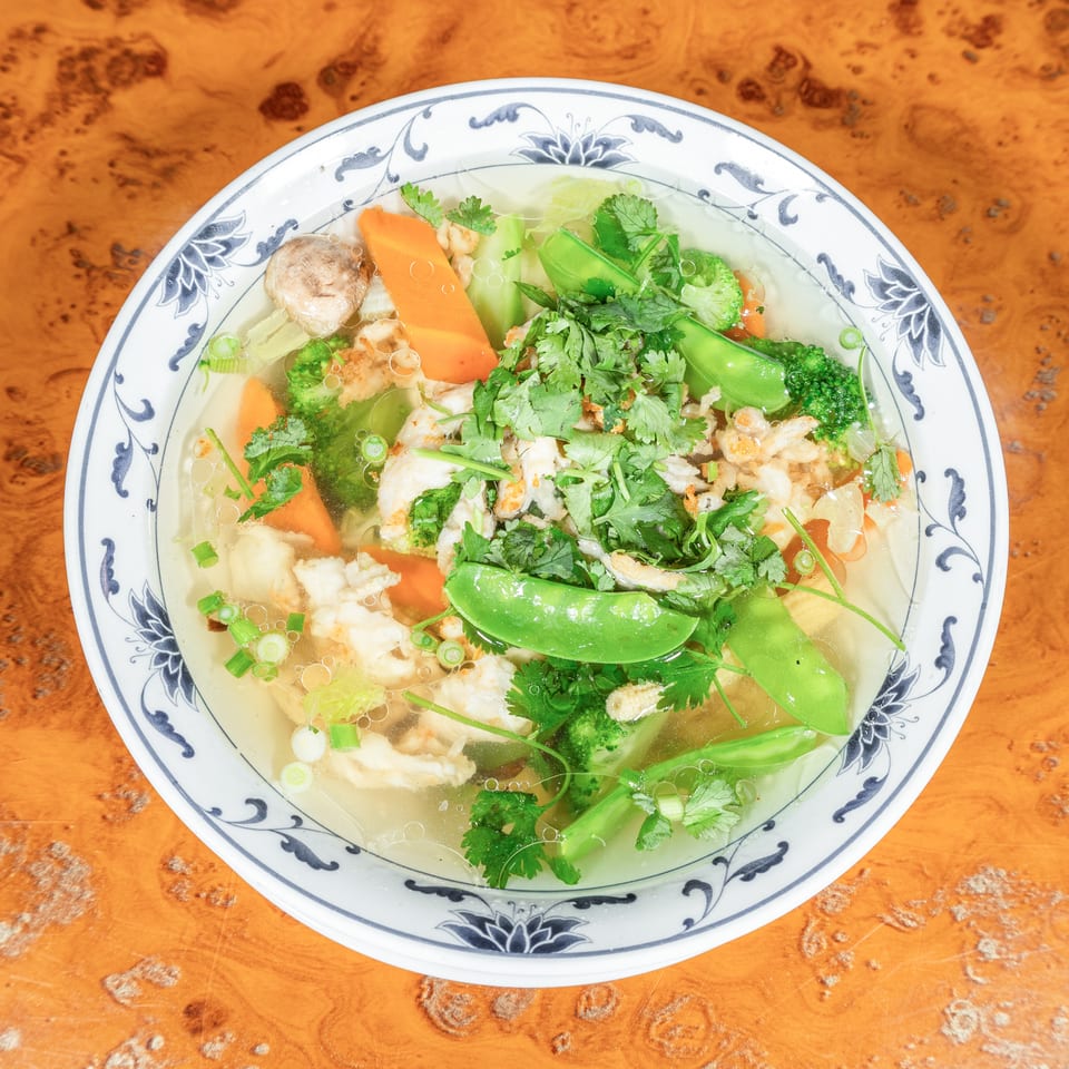Bowl of clear vegetable soup with shrimp, broccoli, snap peas, carrots, mushrooms, cilantro, and scallions on a wooden table.