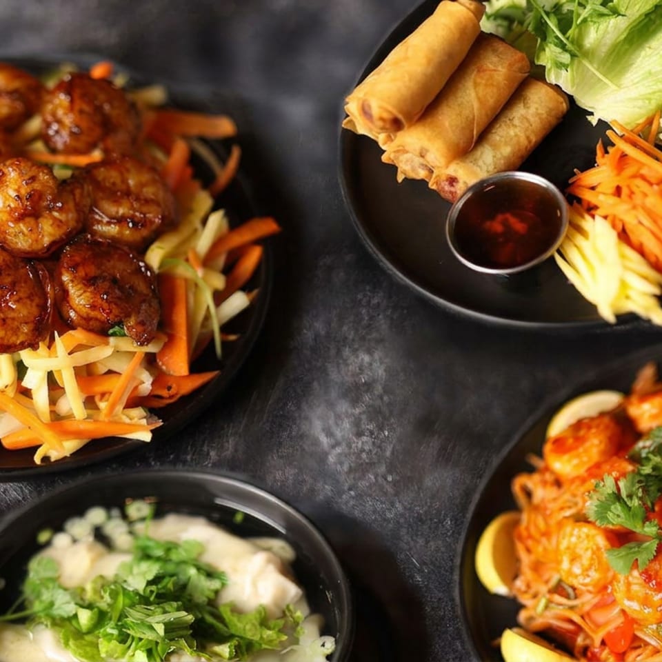 Assorted Asian dishes on black plates, including glazed meatballs over vegetables, spring rolls with fresh garnish, noodles, and dipping sauce.