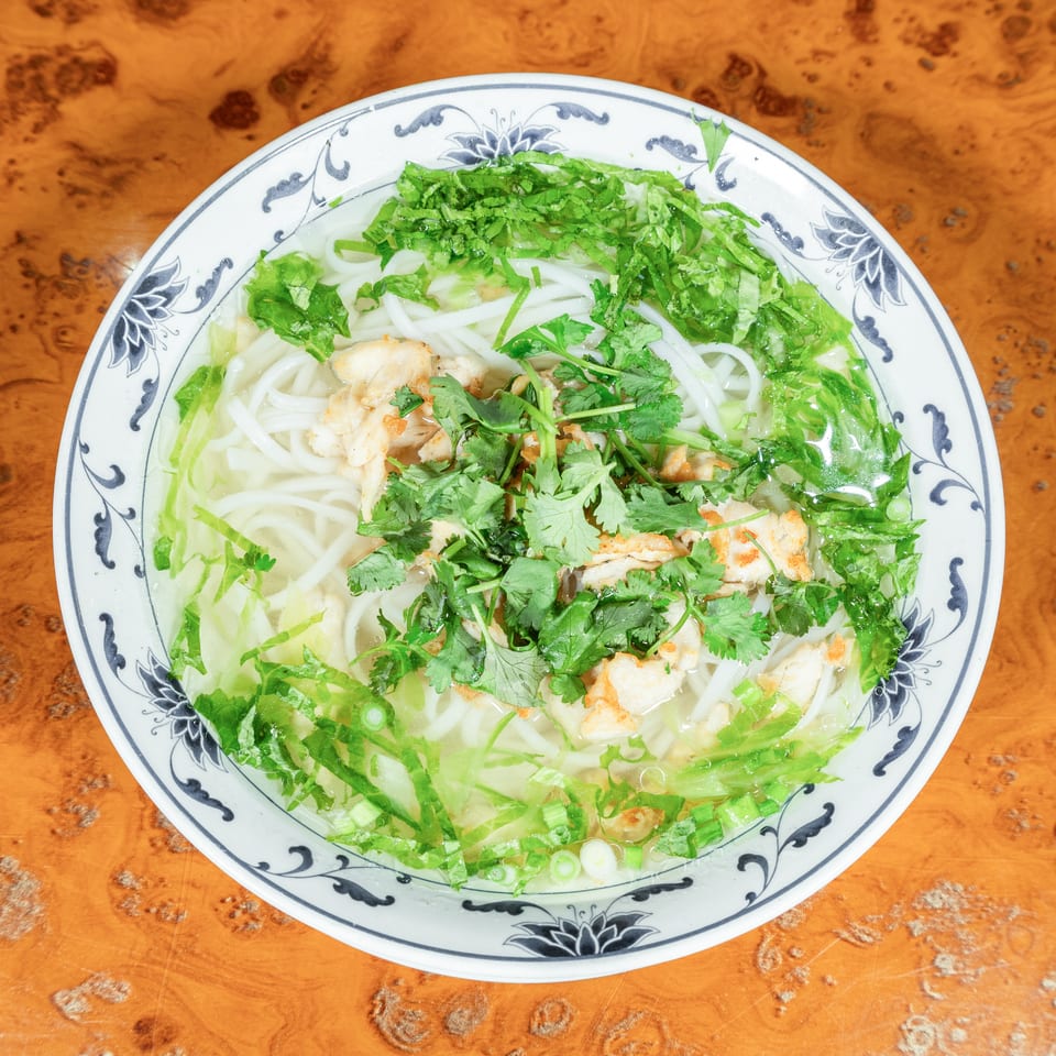 Bowl of rice noodles in clear broth topped with grilled chicken, leafy greens, scallions, and cilantro on a wooden table.