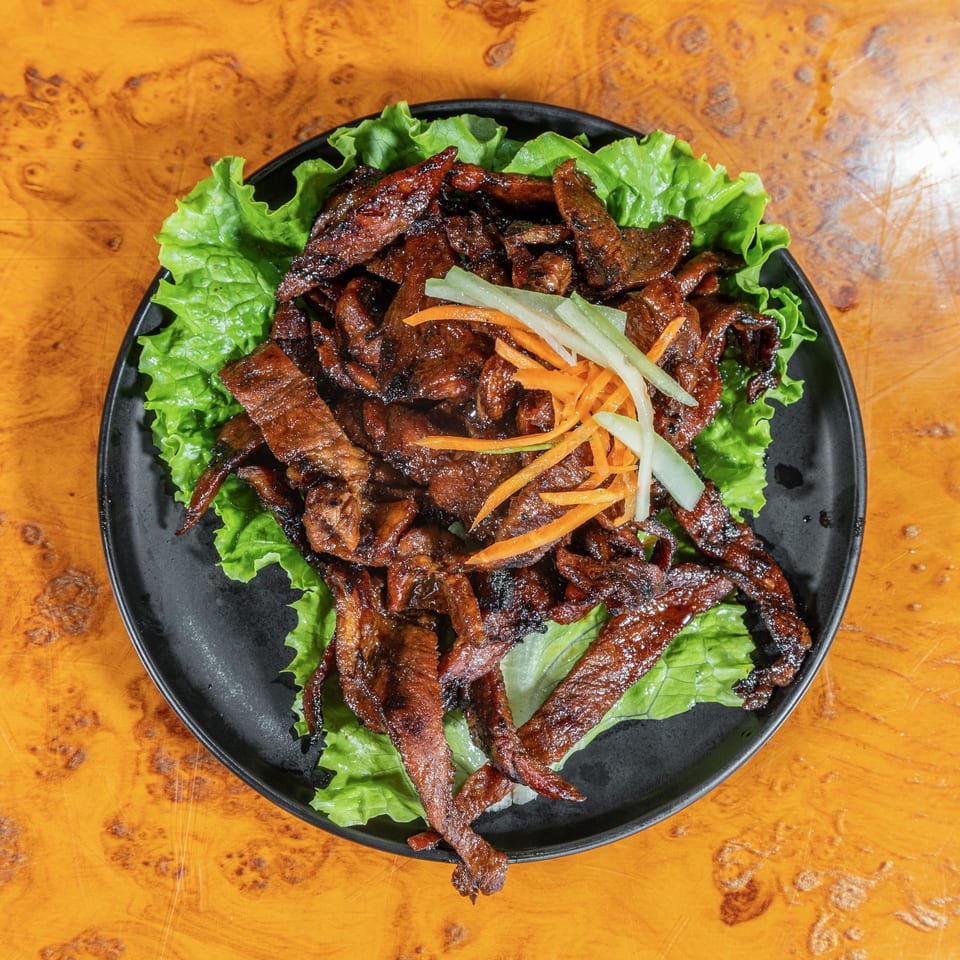 Grilled strips of meat served on lettuce with shredded carrots and cucumber on a black plate atop a wooden table.